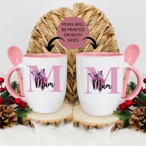 Könnte beinhalten: Zwei rosa und weiße Keramikbecher mit einem Löffelgriff. Jeder Becher zeigt einen rosa Buchstaben "M" mit einem Schmetterlings- und Blumendesign. Der Text "Mum" ist unter dem Buchstaben gedruckt. Die Becher stehen auf einer Holzoberfläche. Die Becher werden auf beiden Seiten bedruckt.