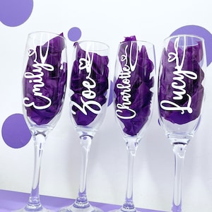 Könnte beinhalten: Vier klare Champagnergläser mit weißer Beschriftung an der Seite. Die Gläser sind mit violettem Seidenpapier gefüllt. Die Namen auf den Gläsern sind Emily, Zoe, Charlotte und Lucy.