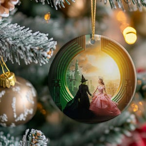 May include: Round Christmas ornament featuring a scene from the Wizard of Oz. The ornament depicts the Wicked Witch of the West and Glinda the Good Witch holding hands. The background shows the Emerald City and a sunset.