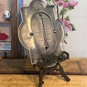 Op de afbeelding: Antiek zilverkleurig thermometer met een zonontwerp en de tekst "Celsius Reaumur Fahrenheit" op het gezicht. De thermometer is gemonteerd op een decoratieve messing standaard.