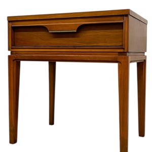 May include: A mid-century modern nightstand with a single drawer and a rectangular top. The nightstand is made of wood with a warm brown finish. It has four slender legs and a metal drawer pull.