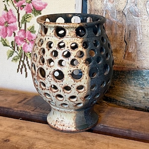 Puede incluir: Un jarrón de cerámica con un esmalte moteado en tonos marrones, blancos y negros. El jarrón tiene un cuerpo redondo con una abertura ancha y una base con un pie pequeño. El jarrón está decorado con un patrón de agujeros.