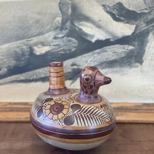 May include: A hand-painted ceramic bird-shaped pitcher with brown, white, and yellow floral designs. The pitcher has a spout and a small handle.