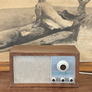 Puede incluir: Una radio vintage de madera con una cara blanca y una perilla plateada. La radio tiene una rejilla de altavoz blanca y una etiqueta que dice "Modelo Veintiuno Cambridge Massachusetts".