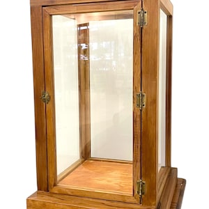 May include: A wooden display cabinet with glass panels and a hinged door. The cabinet is made of light brown wood and sits on a matching wooden stand. The glass panels allow for clear viewing of the interior.