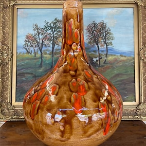 Può includere: Un vaso in ceramica marrone con dettagli arancioni e bianchi. Il vaso ha una base bulbosa e un collo alto e stretto. La glassa ha un effetto gocciolato. Il vaso è esposto davanti a un dipinto di paesaggio incorniciato.