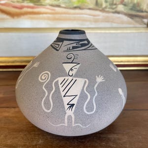 May include: Gray ceramic vase with black and white Southwestern-style designs. The vase features a central figure with outstretched arms and other abstract symbols. The vase has a dark rim and is displayed on a wooden surface.