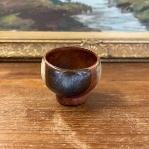 May include: A small, handcrafted ceramic cup with a rounded body and a short, pedestal base. The cup features a colour-blocked design with shades of brown, cream, and dark blue. The interior is a warm brown colour.