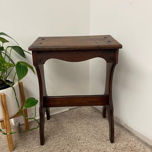 May include: A small, antique wooden side table with a dark brown finish. The table has a rectangular top and curved legs, connected by a lower stretcher. The wood grain is visible, and the table appears to be well-worn.
