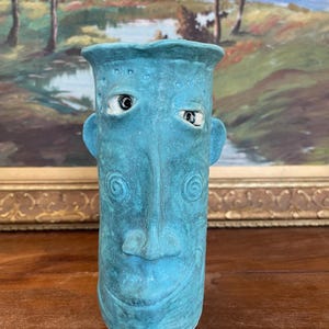 Puede incluir: Un jarrón de cerámica turquesa con diseño de rostro. El jarrón tiene forma cilíndrica con la parte superior ensanchada y pequeñas protuberancias en forma de oreja. La cara presenta ojos con borde blanco, nariz y boca sonriente. Remolinos decorativos en las mejillas.