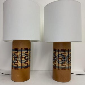 May include: Pair of ceramic table lamps with white lampshades. The lamps feature a brown base with a geometric pattern in shades of blue, brown, and white.
