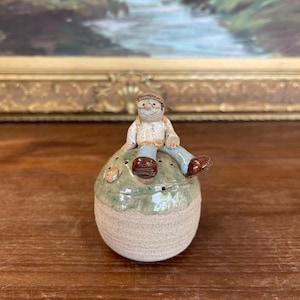May include: A handmade ceramic figurine of a person sitting on a round, light green and beige object. The figurine has a smiling face, wearing a cap, a white shirt, blue trousers, and brown shoes. The object has small holes.