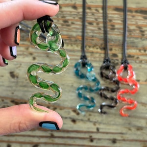 May include: A green and gold glass snake pendant is held up, with three other snake pendants in blue, black, and red in the background. The glass snakes are detailed with a winding design and are attached to black cords.