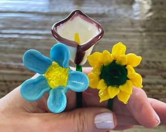 Könnte beinhalten: Eine Hand, die drei Glasblumen hält: eine blaue Blume mit gelbem Zentrum, eine gelbe Sonnenblume und eine weiße Calla mit lila Rand. Jede Blume hat einen grünen Stiel.