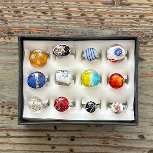 May include: A collection of 12 colorful glass rings displayed in a white box. The rings are made of different colors and patterns, including blue, green, red, yellow, and white.