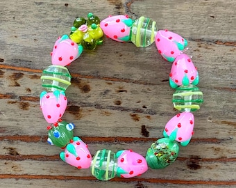 Op de afbeelding: Een roze en groene glazen armband met aardbei-vormige kralen. De kralen zijn versierd met groene bladeren en rode stippen.