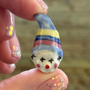 May include: A small, hand-painted ceramic clown figurine. The clown has a white face with black cross-shaped eyes and a red nose. The hat is striped with blue, yellow, and red. The figurine is held between two fingers.