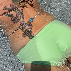 May include: A beaded body chain with a blue evil eye charm. The chain is made of small, colorful beads and is worn over a light green bikini bottom. The water ripples around the body chain.