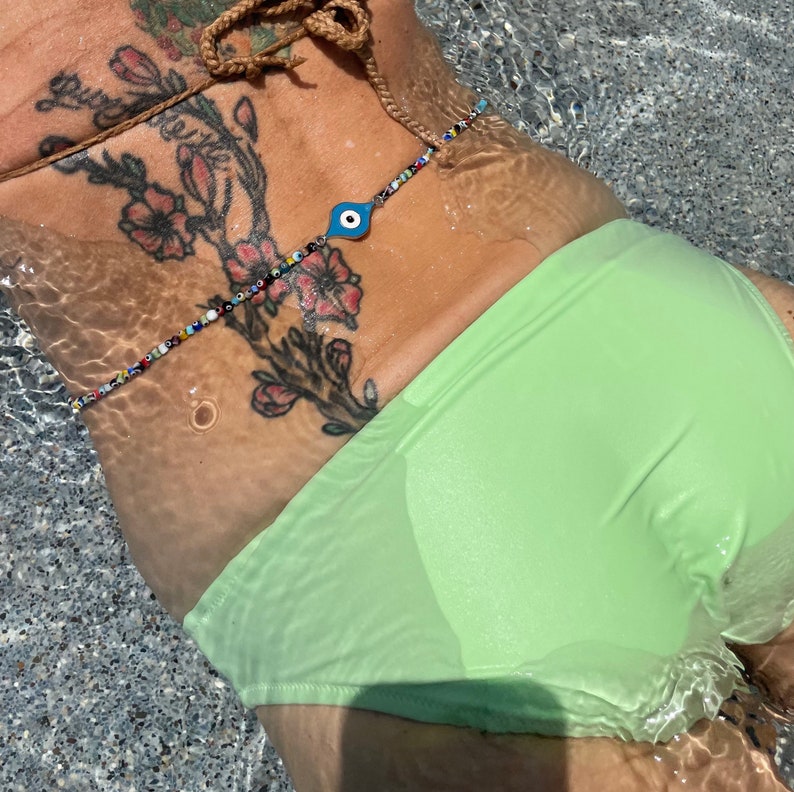 May include: A beaded body chain with a blue evil eye charm. The chain is made of small, colorful beads and is worn over a light green bikini bottom. The water ripples around the body chain.