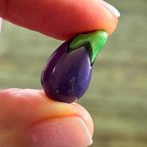 May include: A miniature, glossy purple eggplant with a green stem and black accents. The small, handcrafted vegetable is held between two fingers, showcasing its detailed design and vibrant colors. This decorative piece could be a charm or a small figurine.