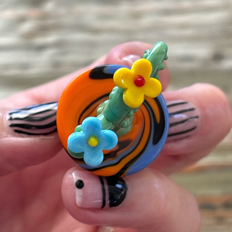 May include: A colorful glass pendant with a swirl design and three flowers. The flowers are blue, yellow, and green. The pendant is on a hand with painted fingernails.
