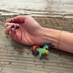 May include: A small glass cactus figurine with a green body, orange and blue flowers, and a striped orange and blue pot. The figurine is sitting on a wooden surface.