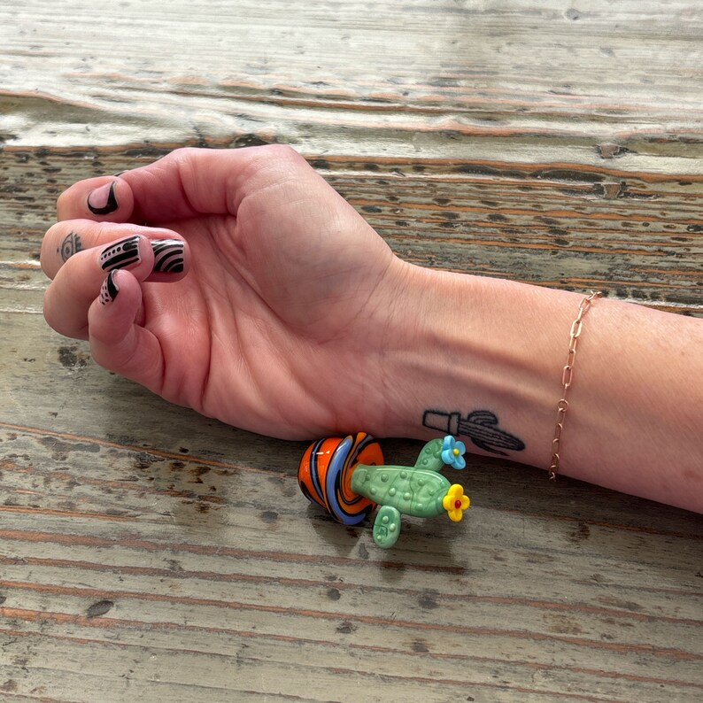 May include: A small glass cactus figurine with a green body, orange and blue flowers, and a striped orange and blue pot. The figurine is sitting on a wooden surface.