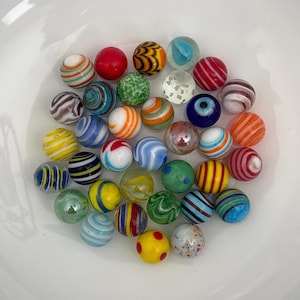 May include: A collection of colorful glass marbles in various patterns and colors, including stripes, swirls, and speckled designs. The marbles are in a white bowl, showcasing a variety of vintage and modern styles.