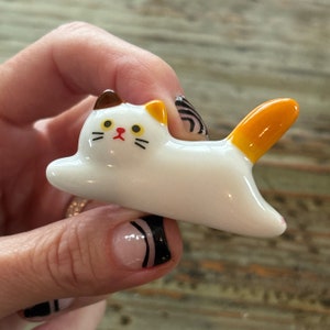May include: A white ceramic cat figurine with orange and brown accents. The cat is lying on its side with its tail curled up.