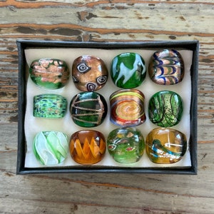 May include: Twelve round glass beads with various patterns and colors, including green, brown, gold, and white. The beads are arranged in a grid pattern in a black box.