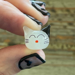 May include: A white glass cat figurine with black ears and whiskers. The cat has a red nose and is smiling.