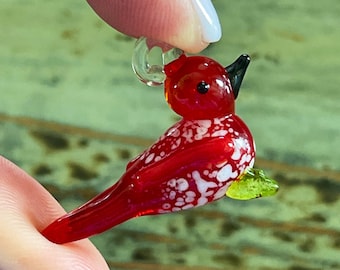 May include: A red glass bird charm with white speckles and a green leaf detail. The bird has a black beak and a clear glass loop for hanging.
