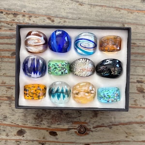 May include: A collection of twelve glass rings arranged in a black display box. The rings feature various designs including swirls, speckles, and solid colors in shades of blue, green, orange, silver, black, and clear glass with embedded patterns.