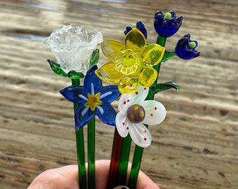 Könnte beinhalten: Ein Strauß aus Glasblumen mit grünen Stielen. Die Blumen sind weiß, blau, gelb und weiß mit roten Punkten. Die Blumen sind in einer Hand angeordnet.