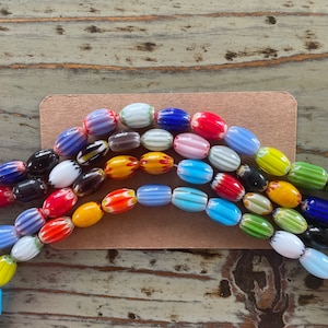 May include: A collection of colorful glass beads with striped and patterned designs. The beads are oval shaped and arranged in a rainbow pattern.
