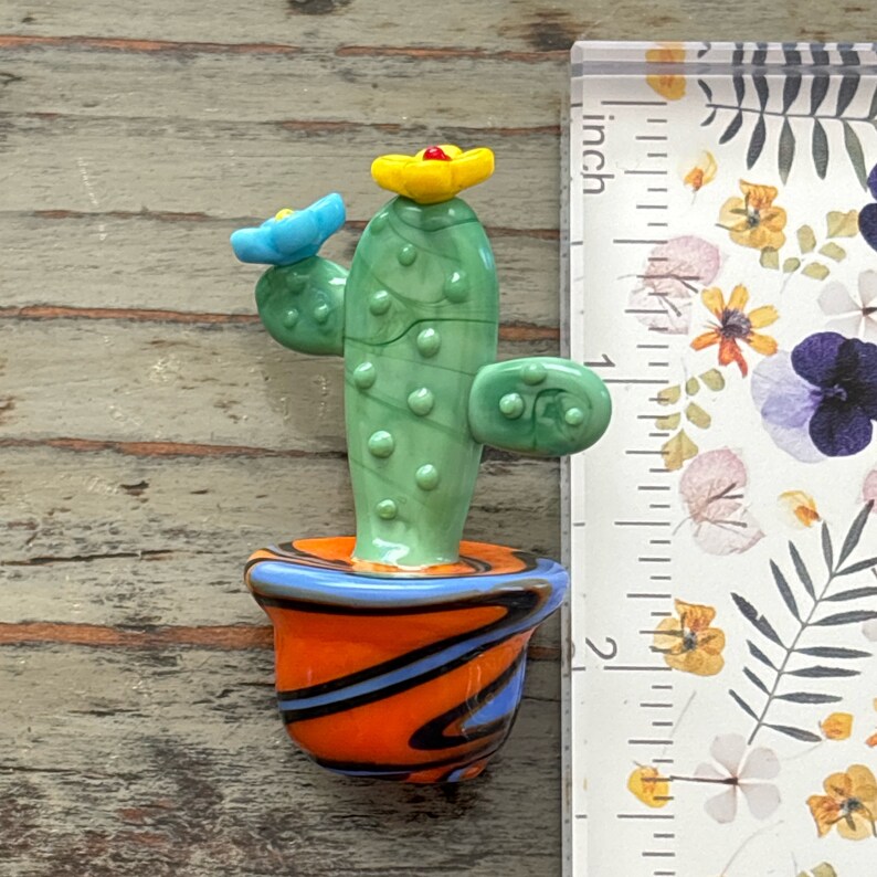 May include: A small glass cactus sculpture with green, blue, and yellow flowers. The cactus is in a small orange, black, and blue swirl pot. The sculpture is about 2 inches tall.