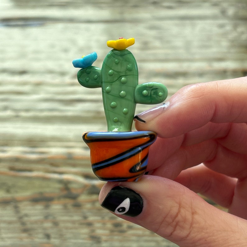 May include: A small green glass cactus with yellow and blue flowers in a colorful orange, blue, and black striped pot.