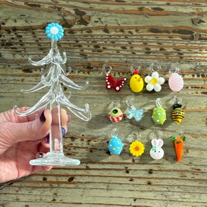 Könnte beinhalten: Ein Glasbaum mit 12 Glasornamenten, die daran hängen. Die Ornamente sind in verschiedenen Farben und Formen, darunter ein Schmetterling, ein Küken, eine Blume, ein Ei, eine Biene, eine Karotte, eine Gänseblümchen und ein Ei. Der Baum steht auf einer Holzoberfläche.