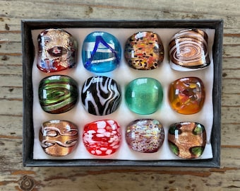 May include: A set of twelve glass rings in a black box. The rings are all different colors and patterns, including green, red, blue, black, white, and gold.