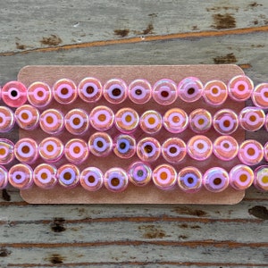 May include: Pink iridescent glass beads with a white and gold circle design. The beads are strung on a string and are displayed on a brown card.