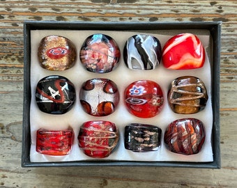 May include: Twelve glass cabochons in a variety of colors and patterns, including red, black, white, and gold, arranged in a display box.