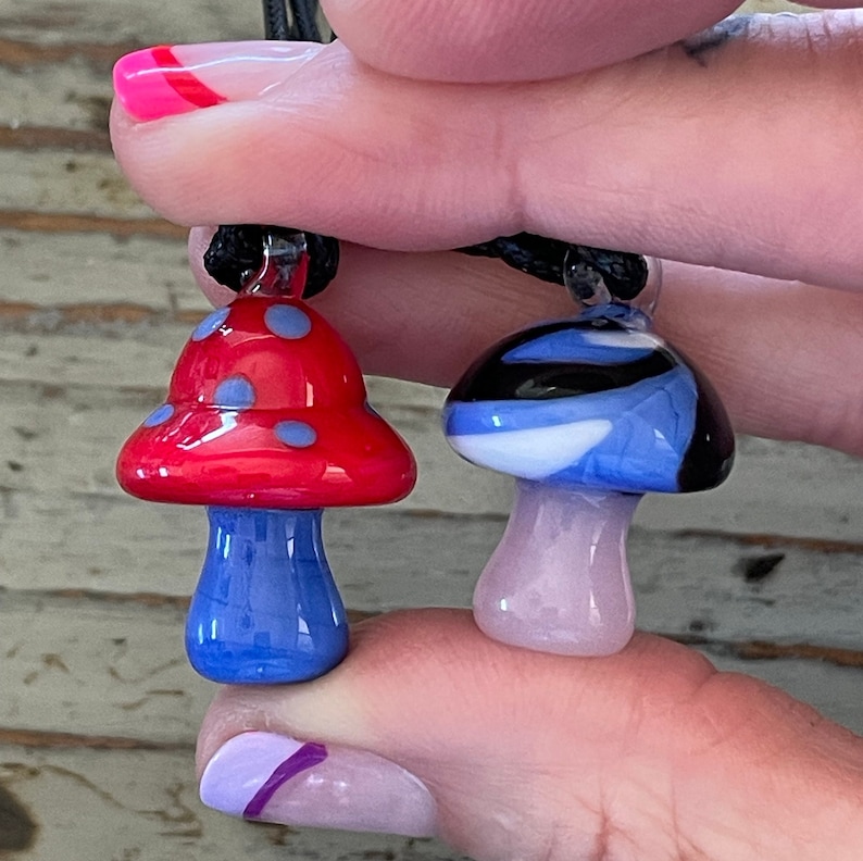 May include: Two glass mushroom pendants on a black cord. One mushroom is red with blue polka dots and a blue stem. The other mushroom is blue with white and black swirls and a pink stem.