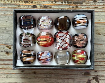 May include: A set of twelve glass rings in a black box. The rings are all different colors and patterns, including brown, white, gold, and red.
