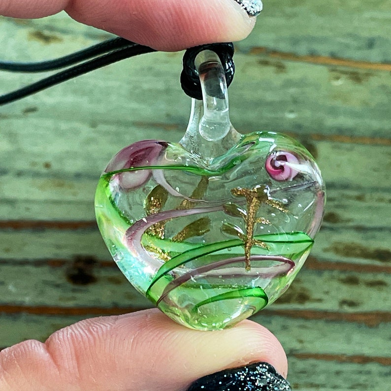 May include: A green glass heart pendant with gold and pink swirls inside. The pendant is on a black cord.
