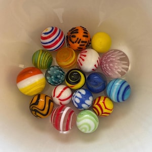Puede incluir: Una colección de canicas de vidrio de colores con una variedad de patrones y diseños. Las canicas presentan rayas, espirales y otros elementos decorativos en tonos de rojo, azul, amarillo, naranja y verde. Las canicas están en un cuenco blanco.