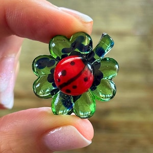 May include: A green four-leaf clover with a red ladybug in the centre. The ladybug has black spots and a black line down the centre.