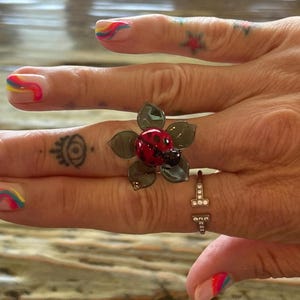 May include: A flower-shaped ring with a red ladybug center and green petals. A second ring with a T-shaped design and small clear stones is also visible. The nails are painted with rainbow designs.