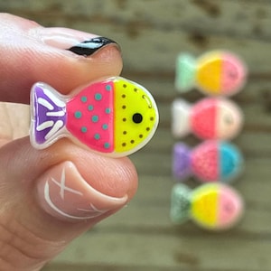 May include: A colorful fish-shaped charm with a pink and yellow body, teal polka dots, and a purple tail fin. The charm is held between fingers, with several similar charms in the background. The fish charm is a vibrant accessory.