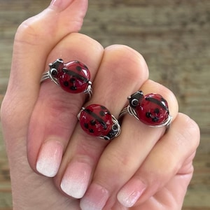 May include: Three ladybug rings with red and black details are displayed. The rings have a silver wire band and a rounded ladybug design. The ladybugs are red with black spots and a black head. The rings are on a hand with a light pink and white ombre manicure.