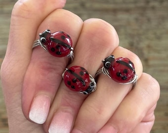 Puede incluir: Se muestran tres anillos de mariquita con detalles rojos y negros. Los anillos tienen una banda de alambre plateado y un diseño de mariquita redondeada. Las mariquitas son rojas con lunares negros y una cabeza negra. Los anillos están en una mano con manicura ombre rosa claro y blanco.